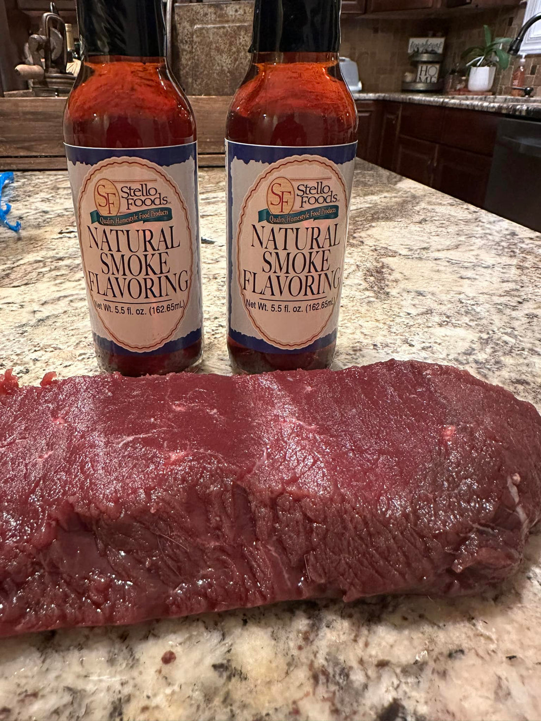 hunk of meat rests on a countertop with two bottles of Stello Foods Natural Smoke Flavoring behind it